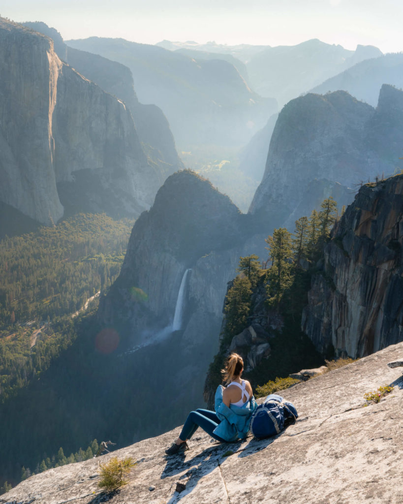 Visiting Yosemite in Spring, Jess sits on a sloped rock face looking across the huge valley and mountains in Yosemite