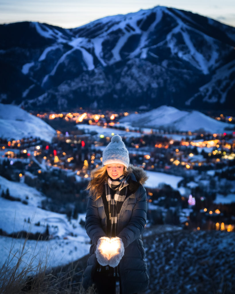 Jess wearing a winter jacket, scarf and hat at the top of a valley overlooking a lit up ski resort at night