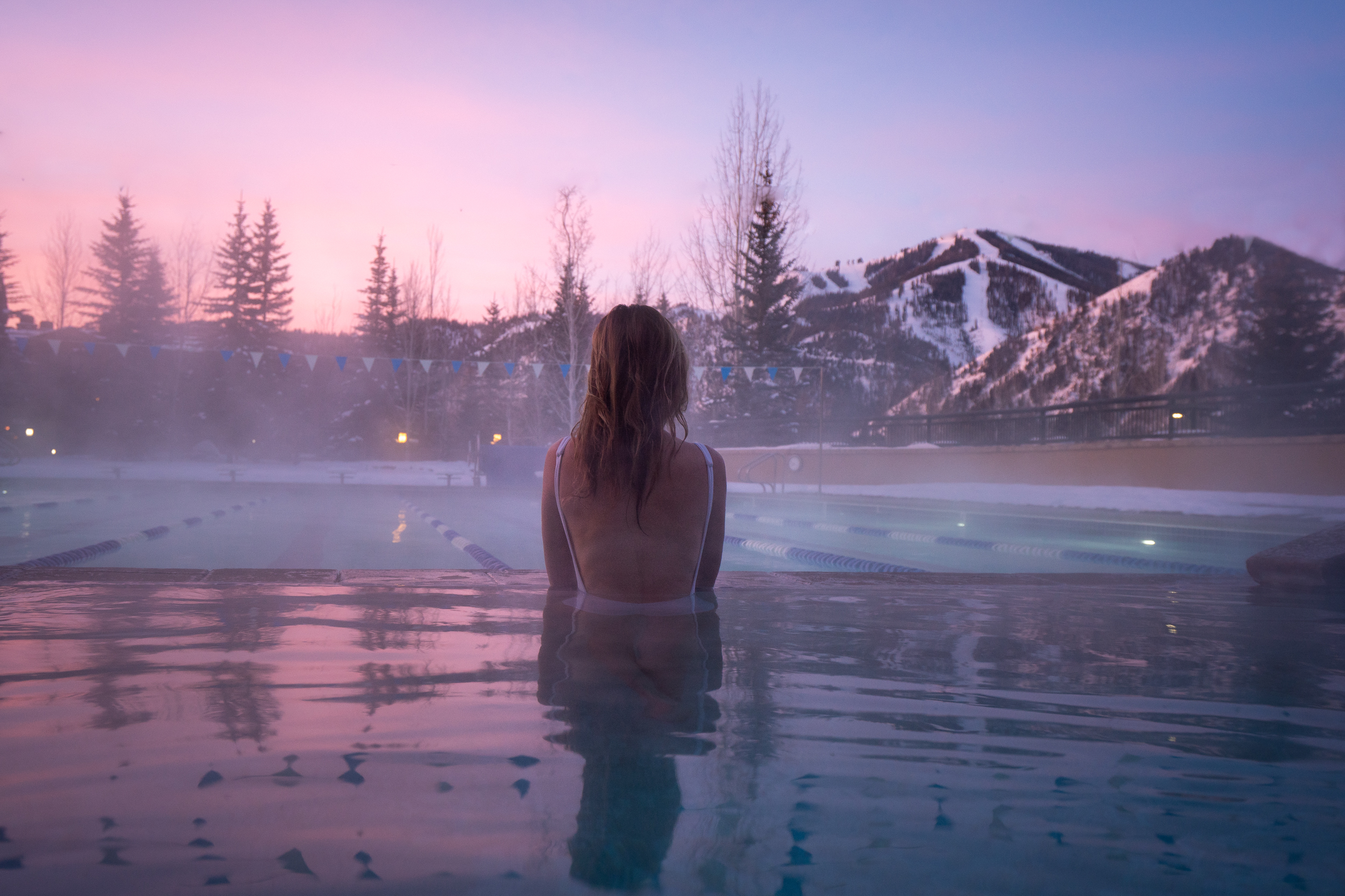 Jess in a steamy spa pool looking across a lap pool with pink sky and snowy mountains in the distance