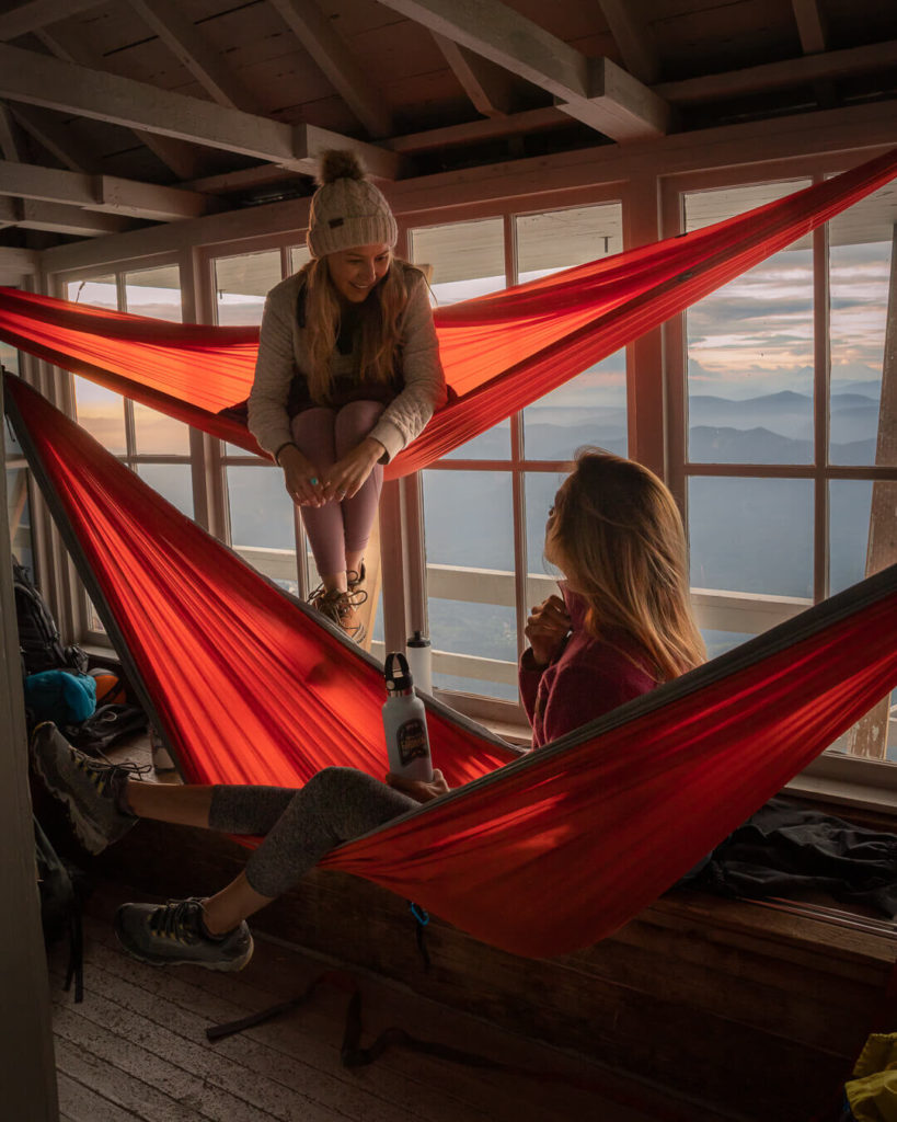 Two women sitting in red hammocks in a fire lookout with mountain views out the window