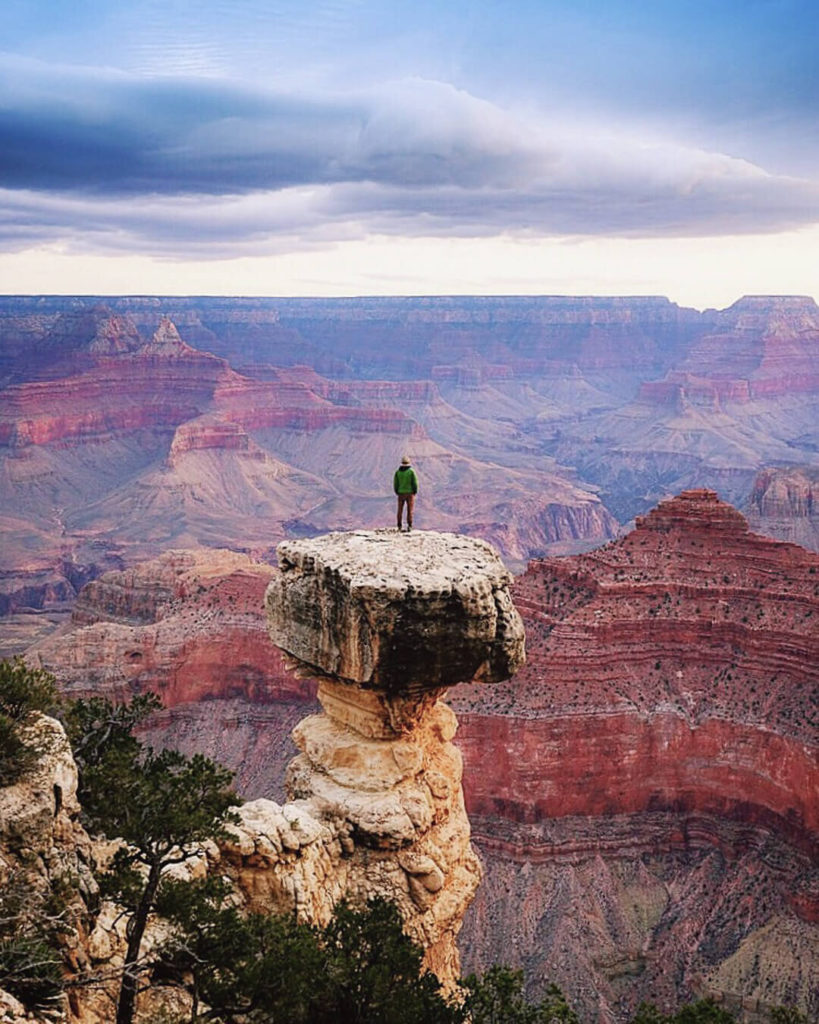A figure in a green coat standing on a rocky pinnacle overlooking the vast red, purple landscape of the Grand Canyon.