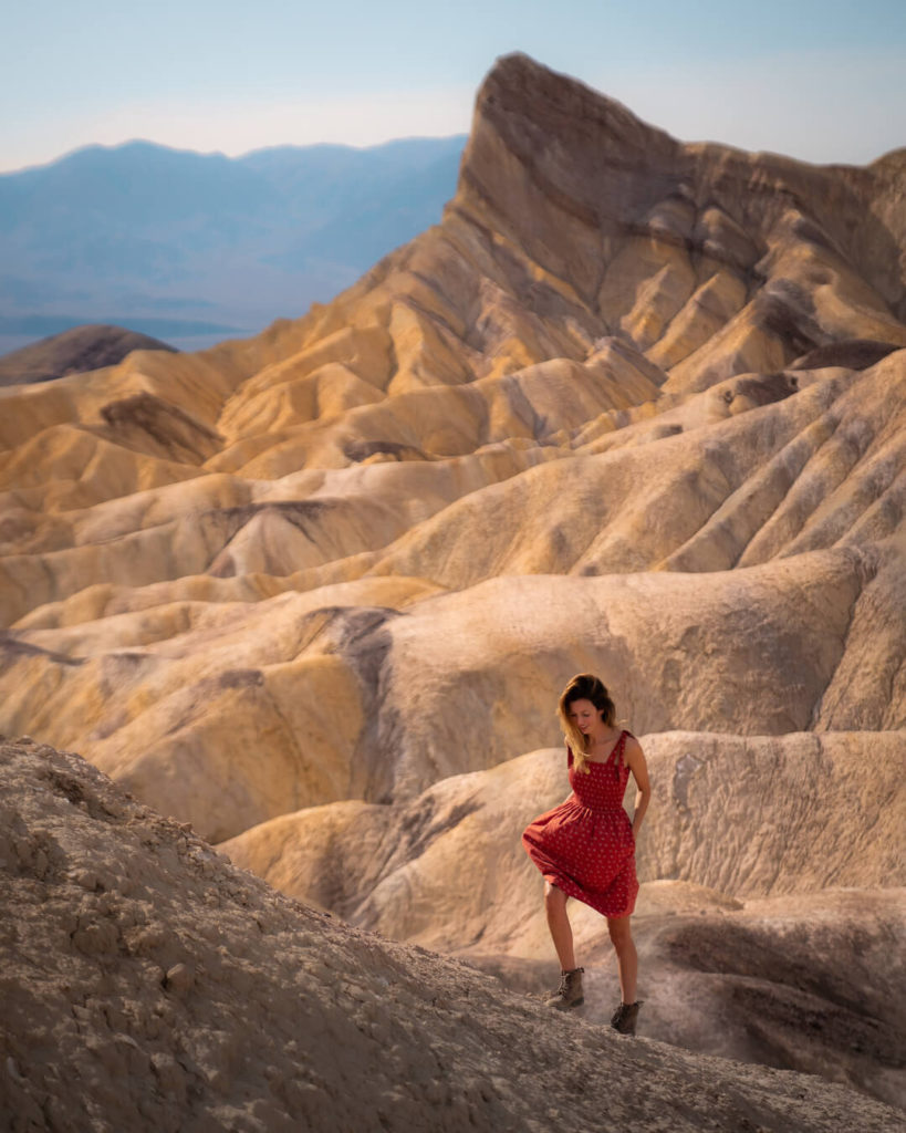 Jess walking up a steep rocky hill in a red dress with a sand-colored barren rocky mountainous landscape behind her.