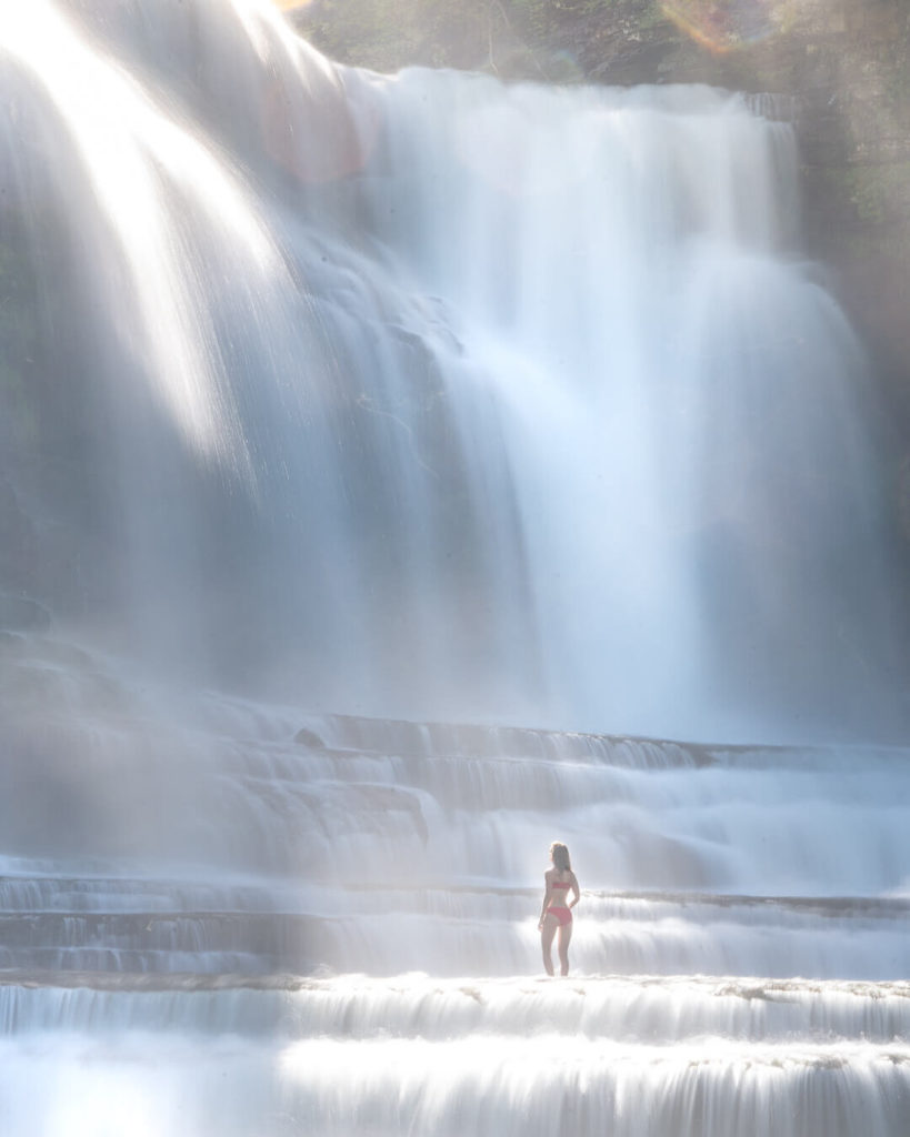 A large waterfall flowing down several tiers of rocks with a figure in a red bathing suit standing in the middle of the rocks. Water is flowing heavily as it's a springtime