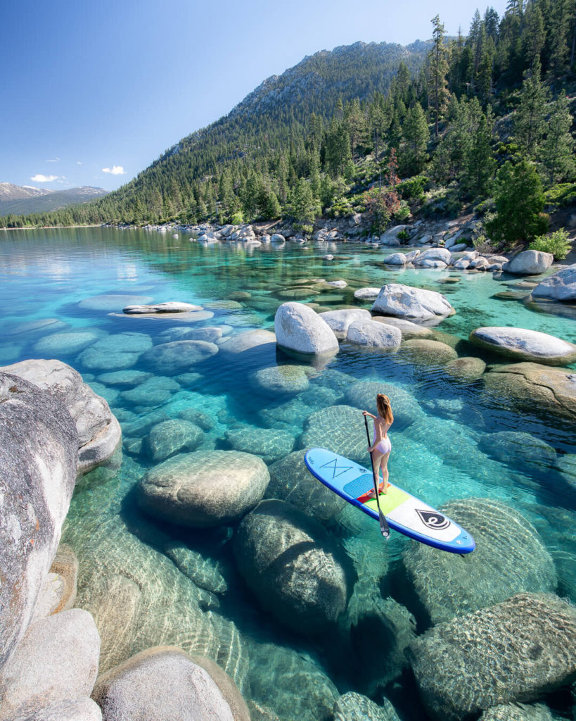 A figure on a stand up paddleboard on the water which is clear blue with large boulders visible in the water at Lake Tahoe.