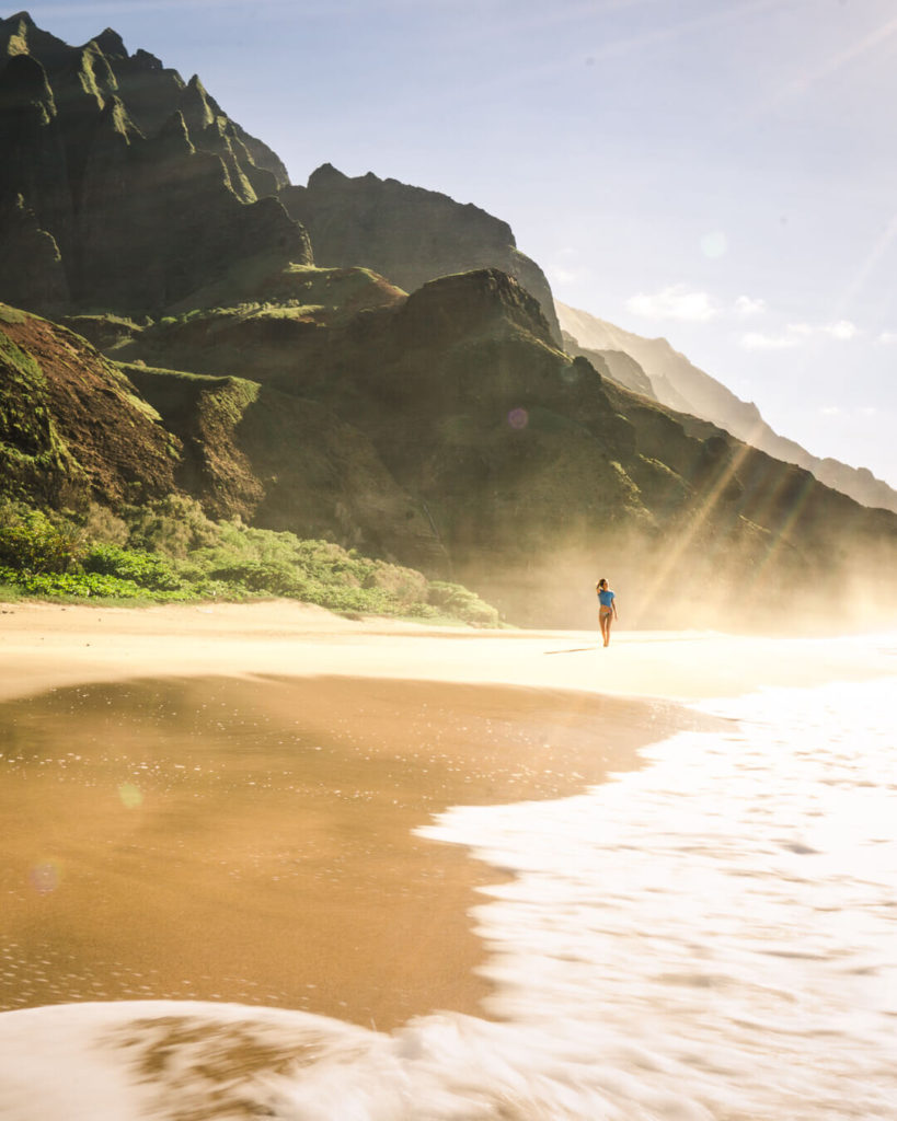 A sandy beach with green mountains behind it and a figure walking along the shoreline in bright sunlight