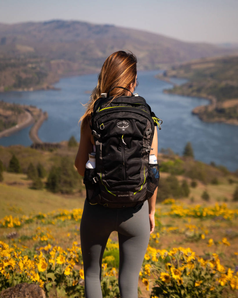 A woman facing away from the camera with a black backpack on her back, looking out across a river