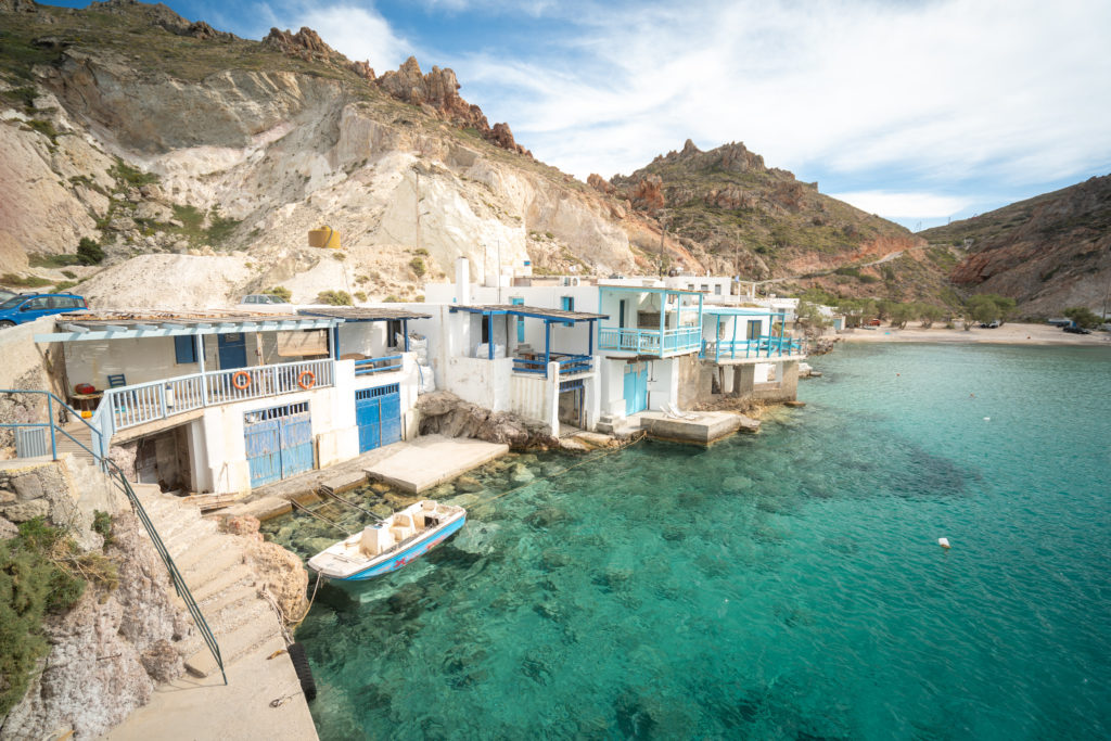 Charming white washed houses along a rocky shore with shallow, turquoise waters. A boat is docked and the doors are painted blue on the houses.