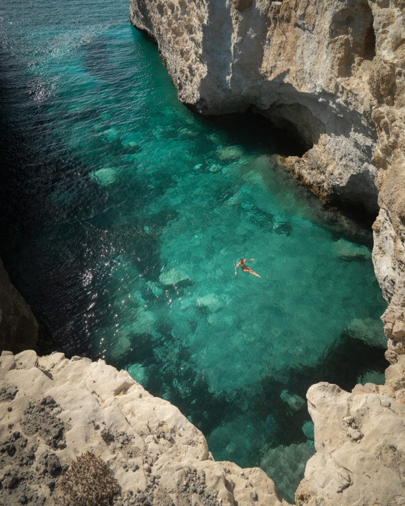 An aerial view of a dramatic rocky cove with turquoise water and a figure swimming in the cove