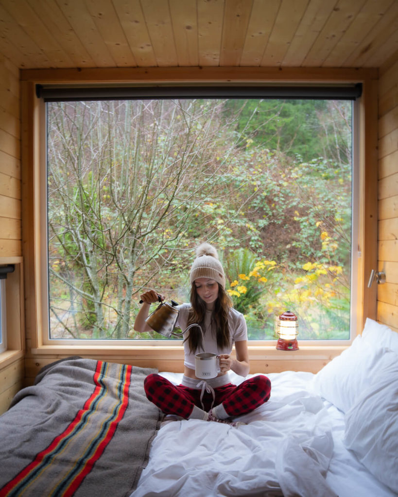 Jess sitting on a bed in a wooden cabin with a kettle, yeti mug and camp lantern