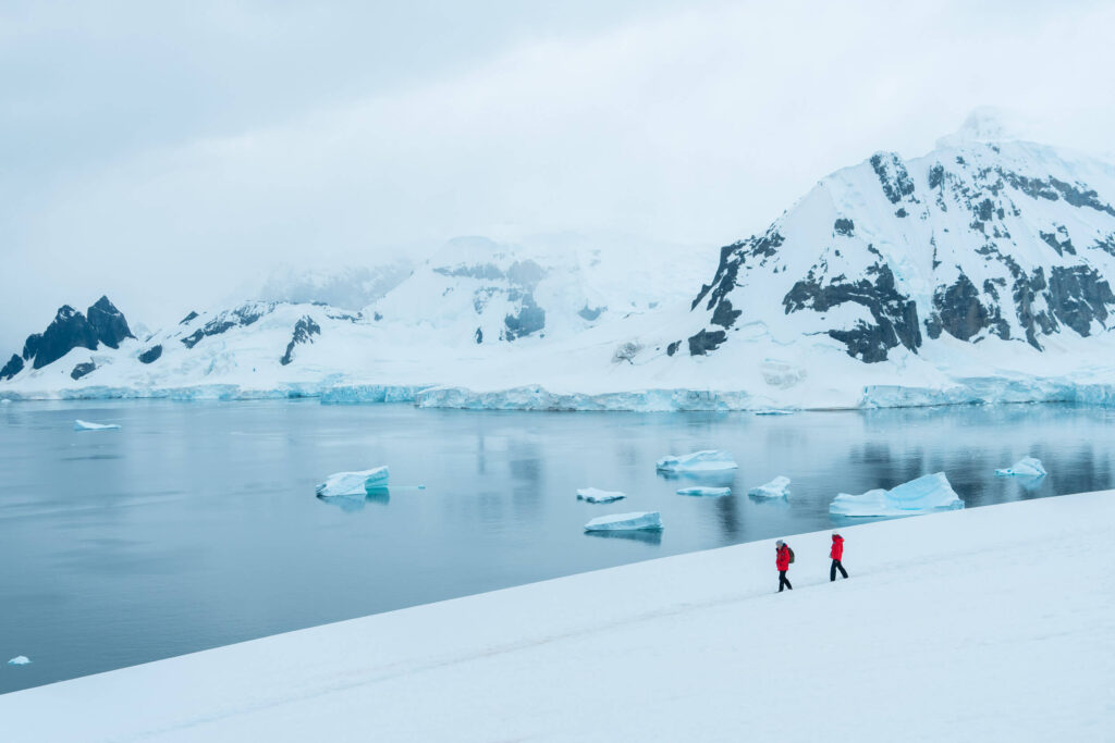 a cloudy day in Antarctica with snow covered mountains, floating icebergs and two people in red coats crossing the snowy landscape in the foreground