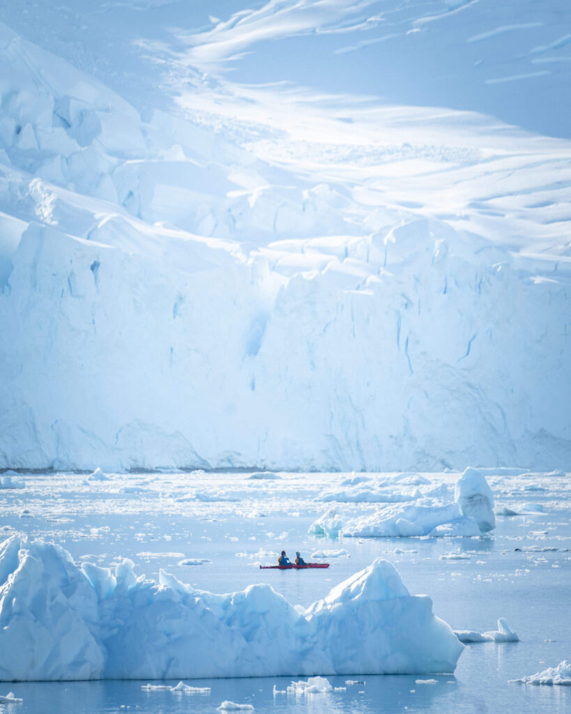 two people in a sea kayak in the distance surrounded by huge ice bergs