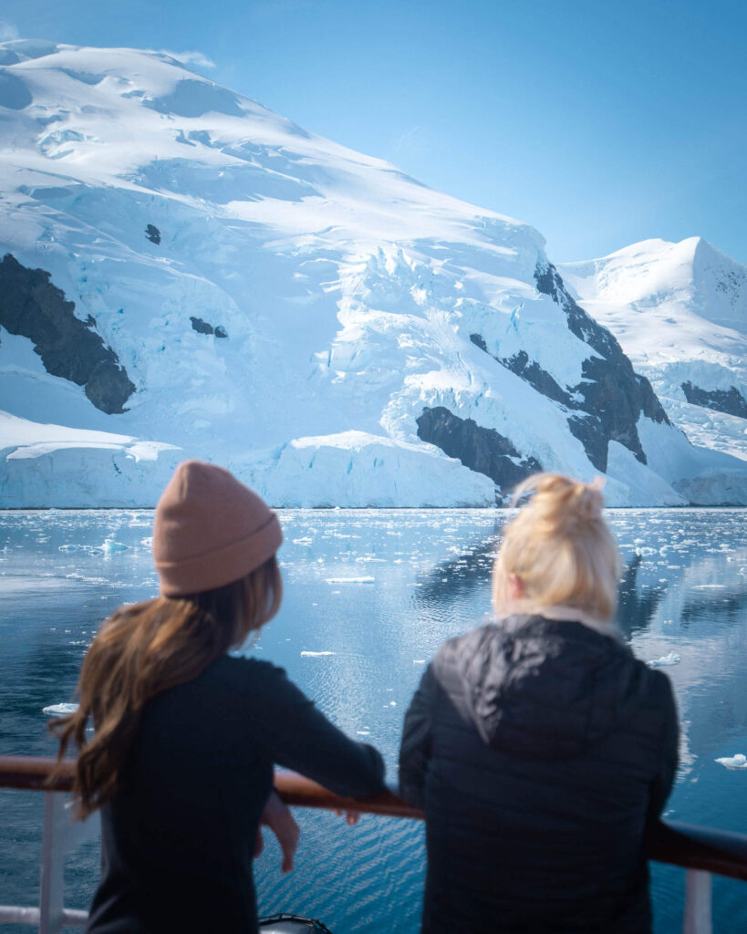 two people looking out from the deck of a cruise ship across the landscape of Antarctica with snow covered mountains and icebergs floating in the water