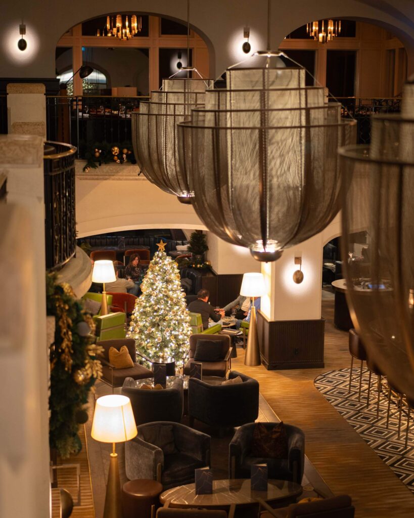 Inside a luxury hotel with large chandeliers and overlooking a balcony where seating areas and a brightly lit Christmas tree can be seen below