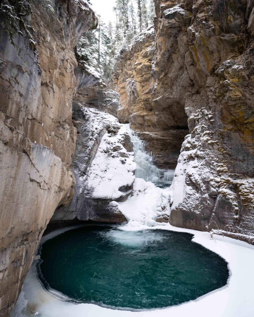 A rocky canyon covered in snow with a dark green pool of water at the bottom which is partially frozen