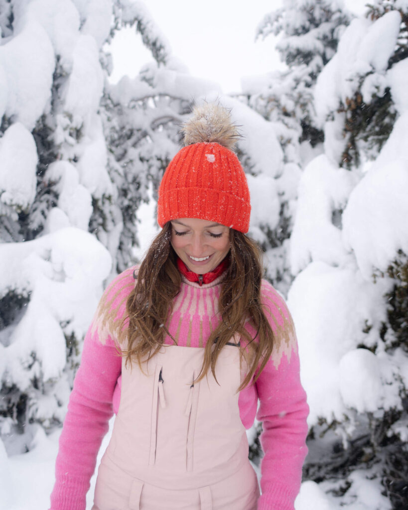 Jess standing in front of snow-covered trees wearing a red hat, pink sweater and pink ski salopettes