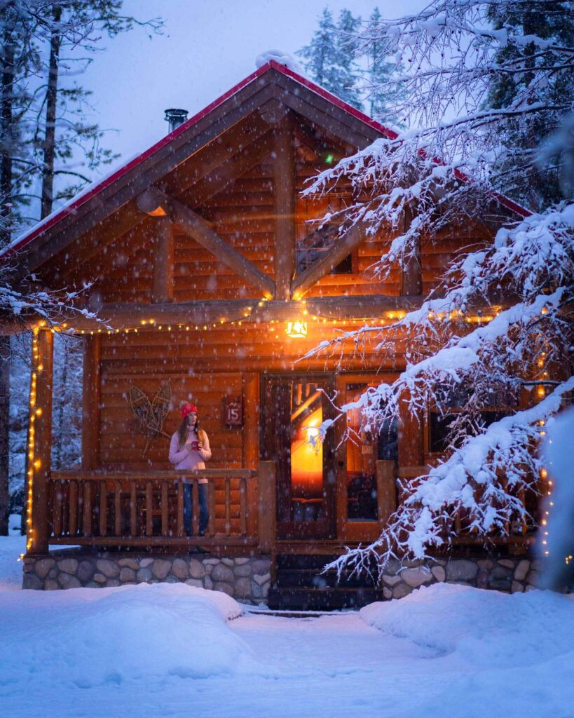 Jess standing on a deck of a log cabin as it snows. She is holding a mug and inside is warmly lit.