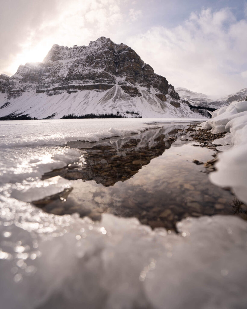 A low-to-the-ground photo of a partially frozen lake with a snowy mountain in the distance