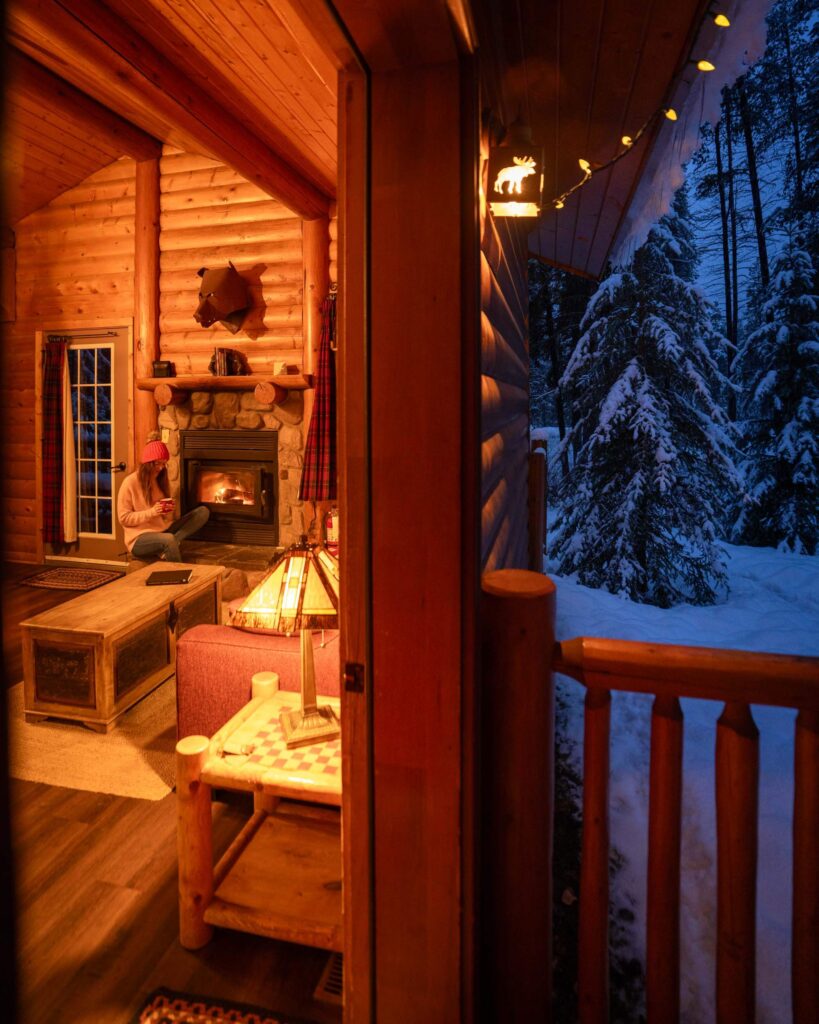 A photo looking through a door frame, outside is dark and snowy, inside is warmly lit and Jess sits by the burning fireplace.