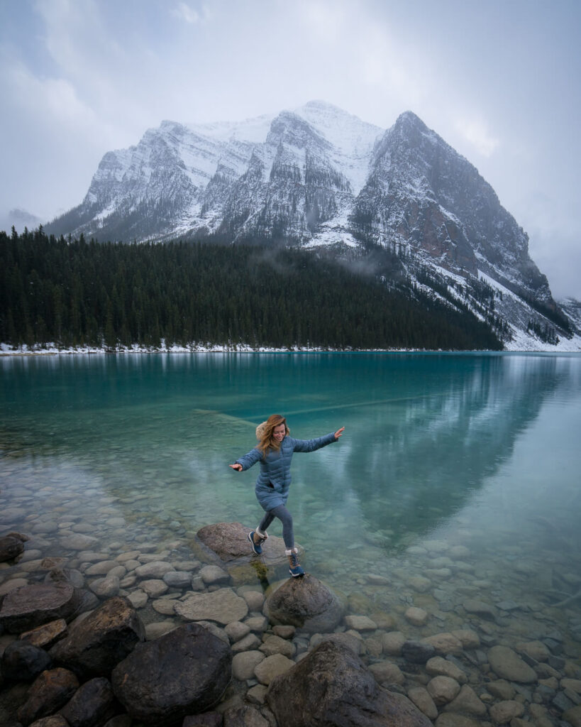 Jess jumping between rocks on the shores of Lake Louise. The water is turquoise blue and there is a snow covered mountain on the far shore