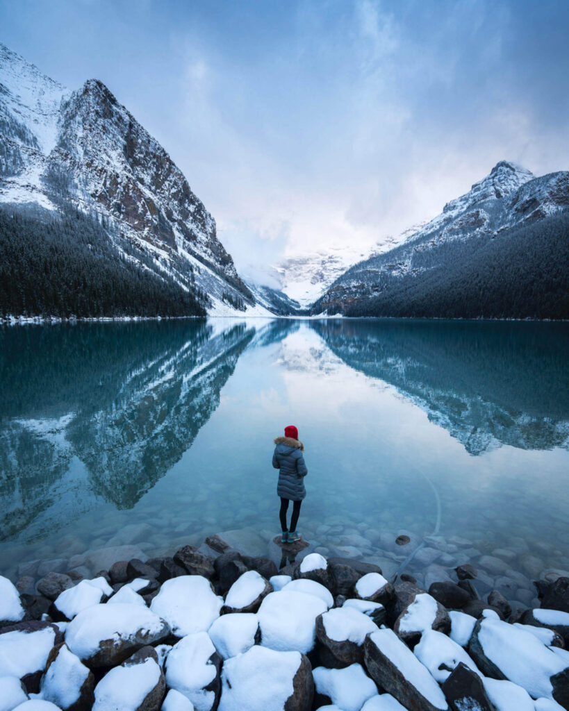 A woman standing on a rock on the edge of Lake Louise looking across the lake. A huge mountain either side is reflected in the water in Banff National Park
