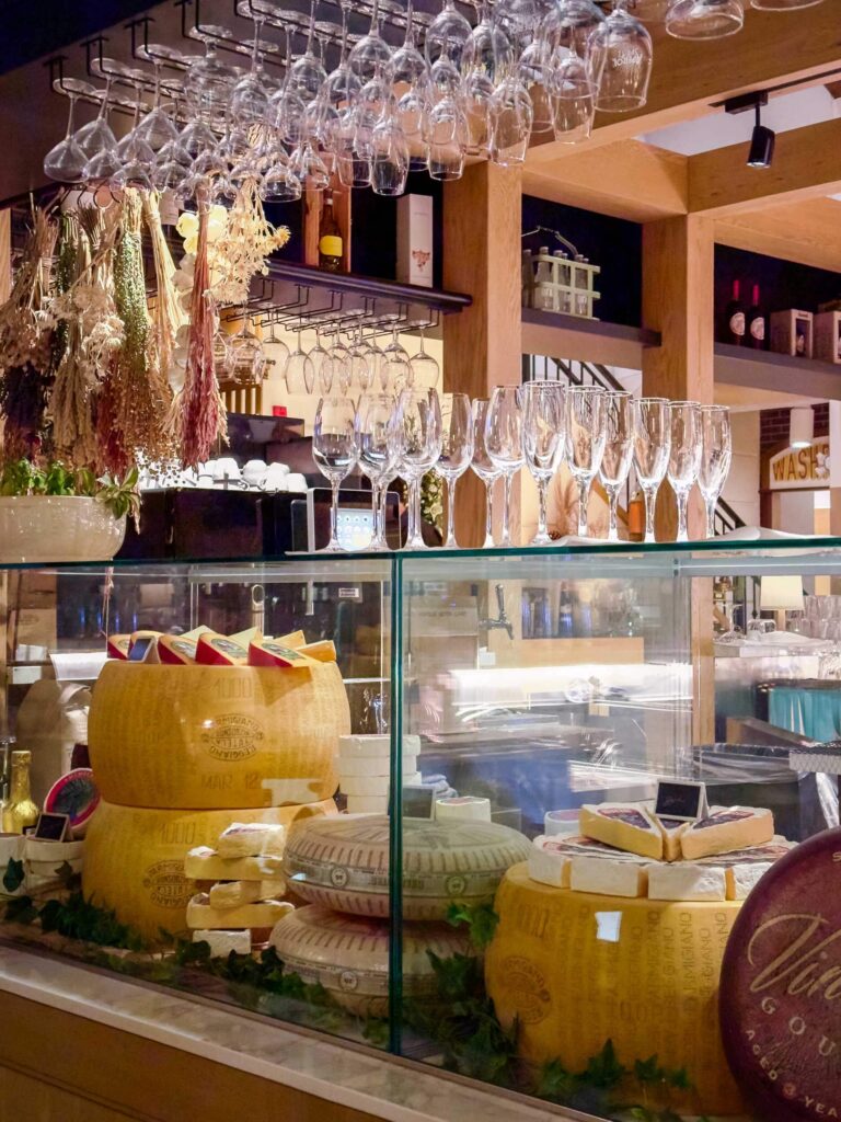 A deli counter with lots of cheeses and wine glasses hanging from the ceiling