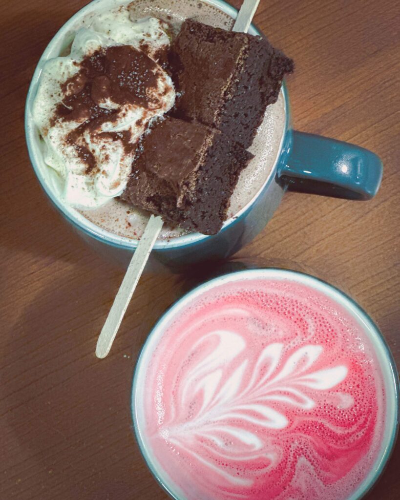 A cup of hot chocolate with a brownie stick on top and a pink cup of beet latte
