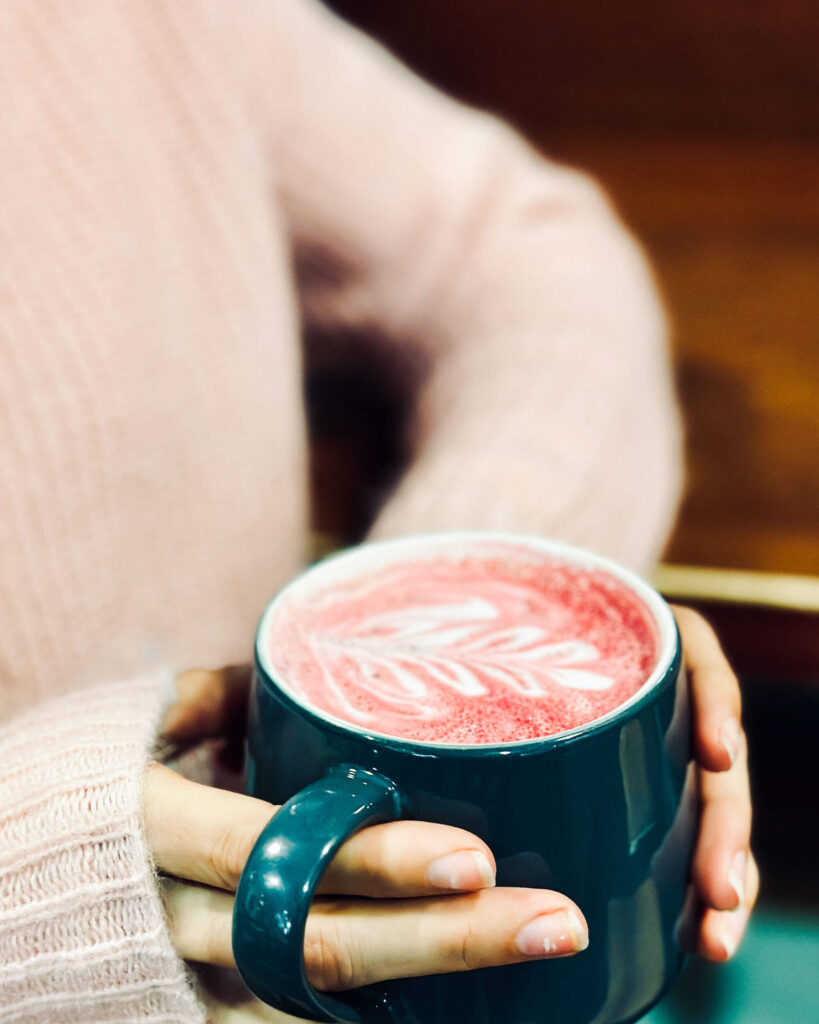 A beet latte with a coffee decoration and in a dark green mug with hands holding it
