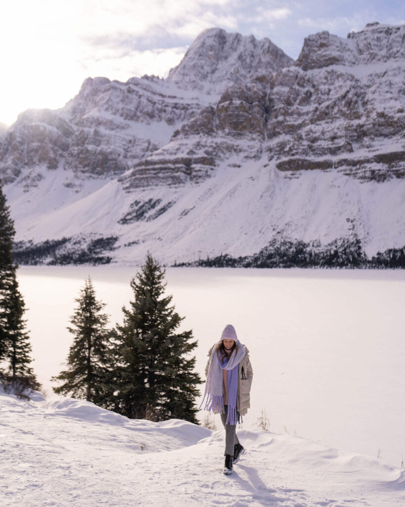 Jess walking on the snow-covered banks of a lake with a snow-covered mountain on the far side. Three spruce trees next to her