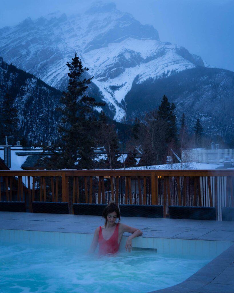Jess in a red swimsuit sitting in a steaming hot tub at a hotel at dusk. Snow covered mountains behind her.