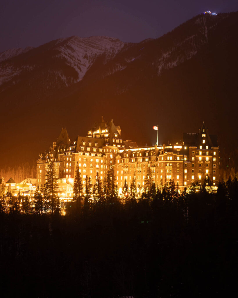 A large, brightly lit hotel building with turrets at dusk, backed by a snowy mountain.