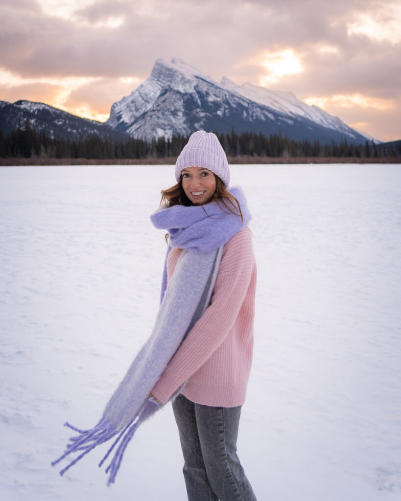 Jess standing on a snow covered frozen lake at sunrise, wearing a pink sweater and purple scarf - one of my favorite Banff winter activities
