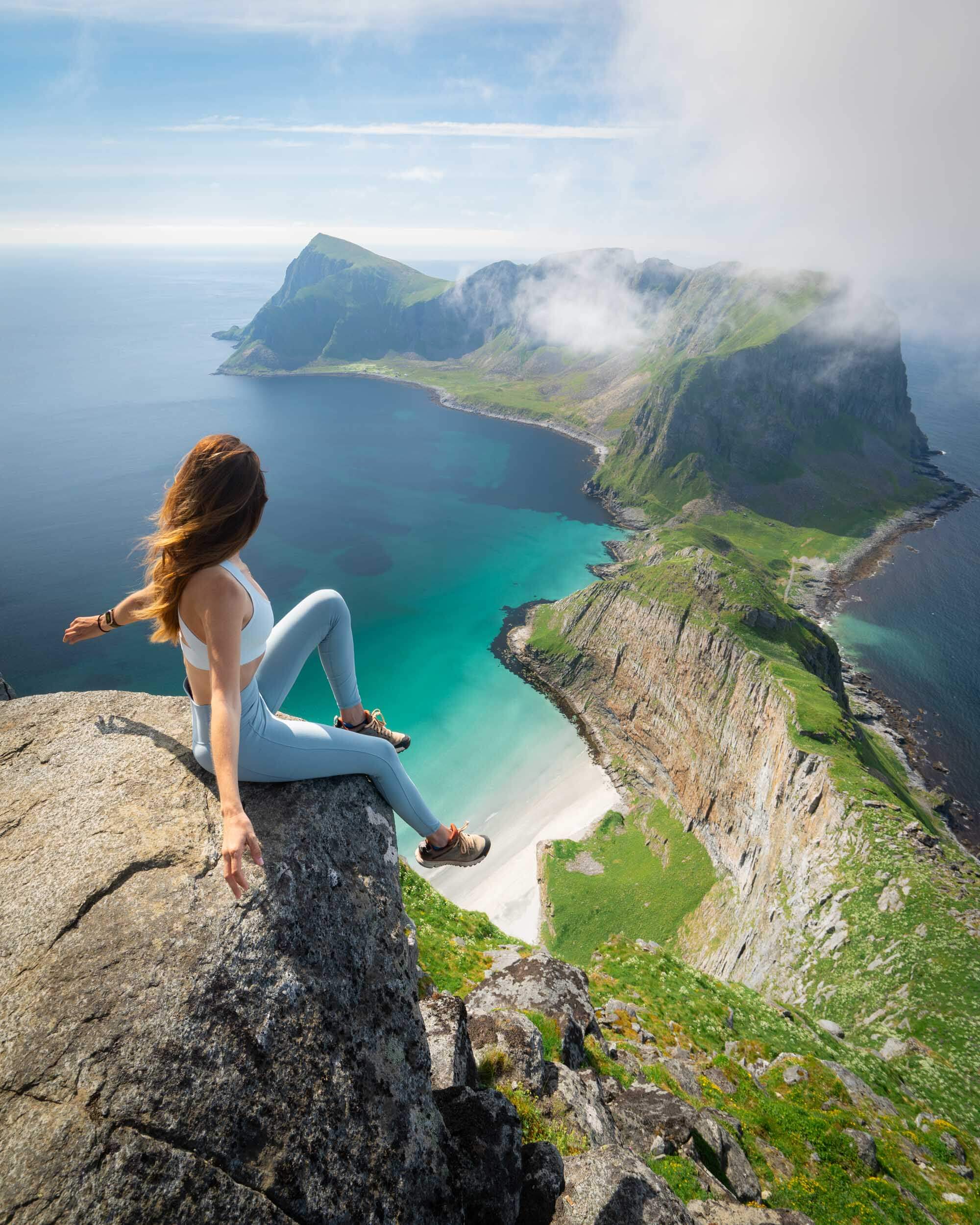 Jess wandering sitting on a rock with arms outstretched with green mountains and blue water on the haen hike on Værøy island in norway