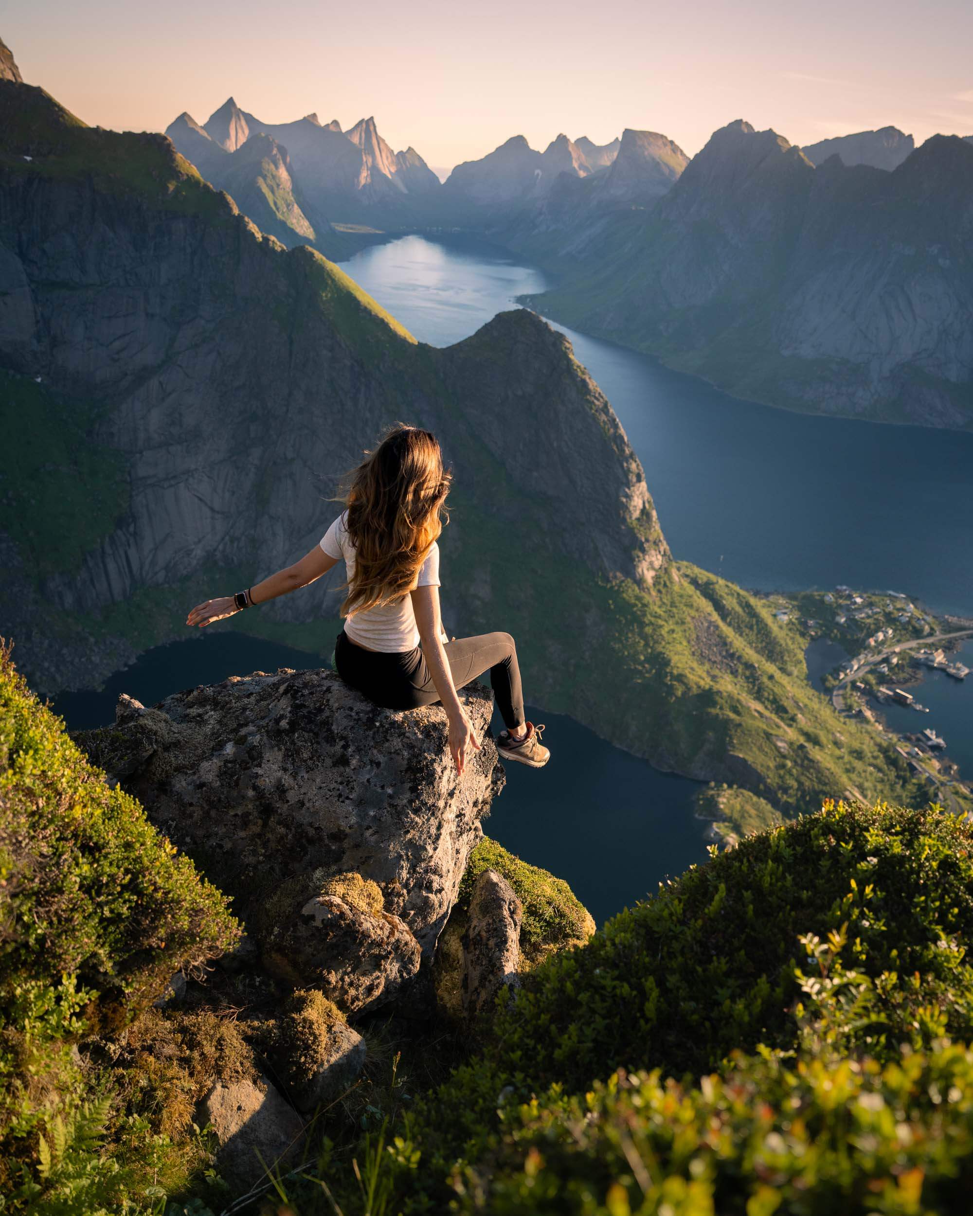 jess wandering sitting with arms outstretched with the view of the mountains and water in the background on the reinebringen hike, one of the best hikes in norway